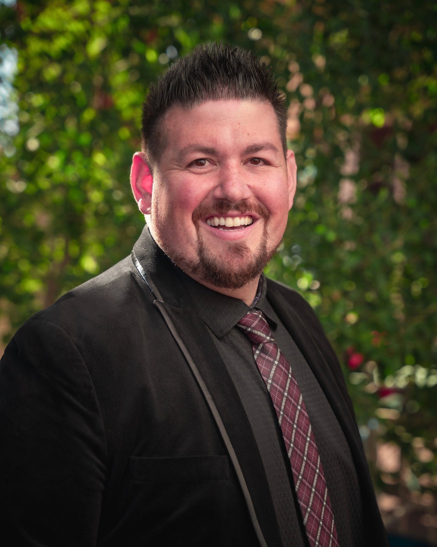 Man in black blazer and patterned tie smiling outdoors with greenery in the background.