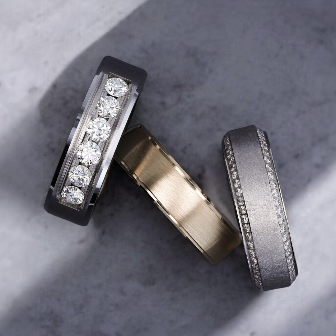 Three men's rings with distinct finishes, featuring channel-set diamonds and brushed metal surfaces, displayed on a stone background for personalized consultation.