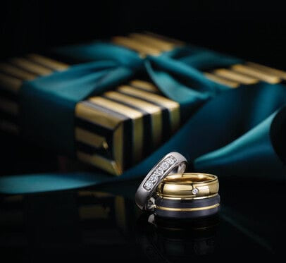 Two gold wedding rings with silver accents on a reflective surface, with a dark background and blue ribbon.