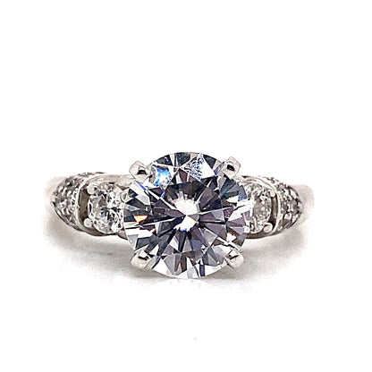 A breathtaking engagement ring showcasing a 6.5 mm round brilliant CZ center, flanked by .70 ct of natural round brilliant diamond accents in a timeless three-stone vintage design, all elegantly set in 14K white gold.