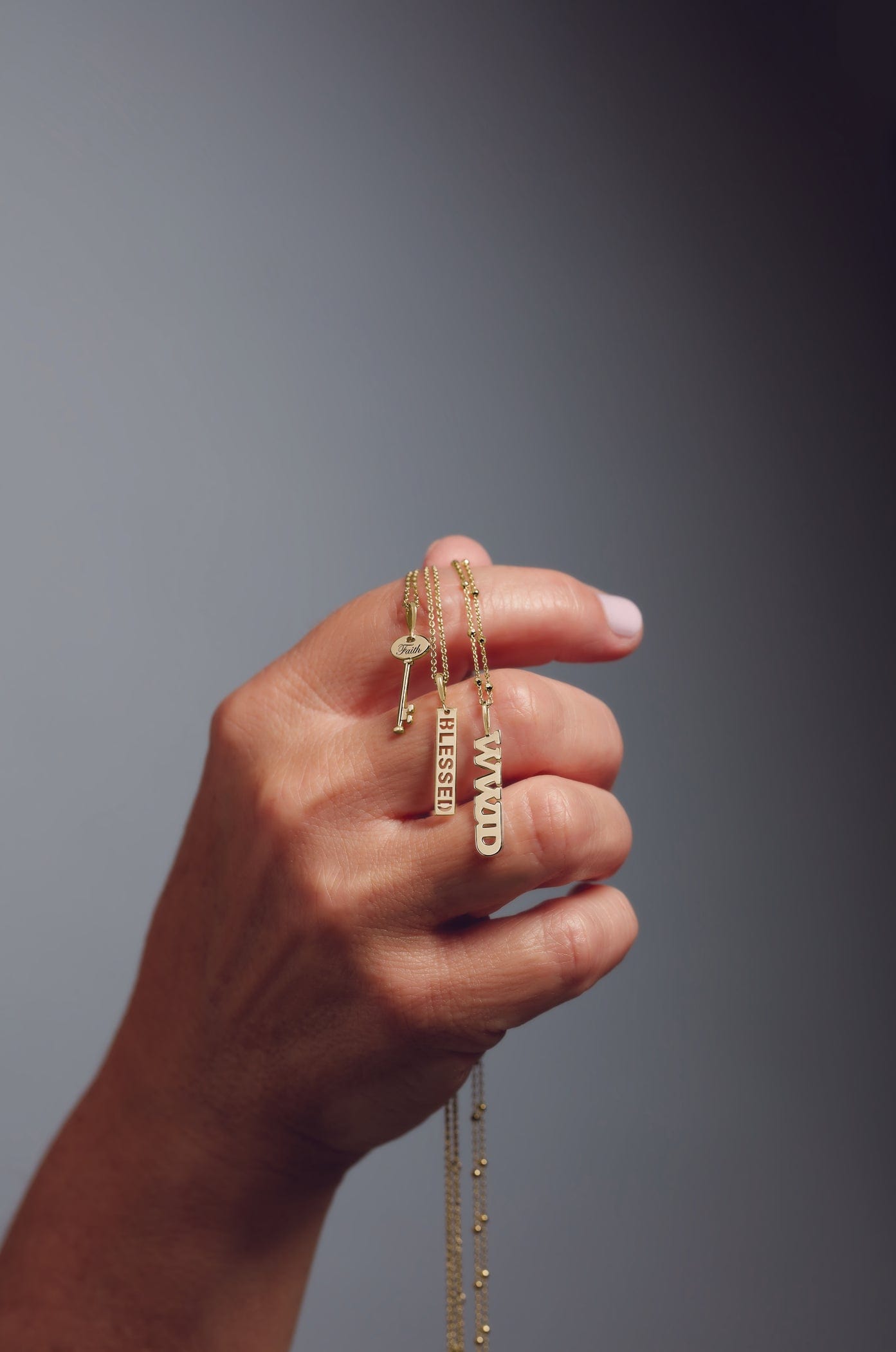 A hand holding gold necklaces with pendant charms spelling BLESSED and MAMA against a gray background.