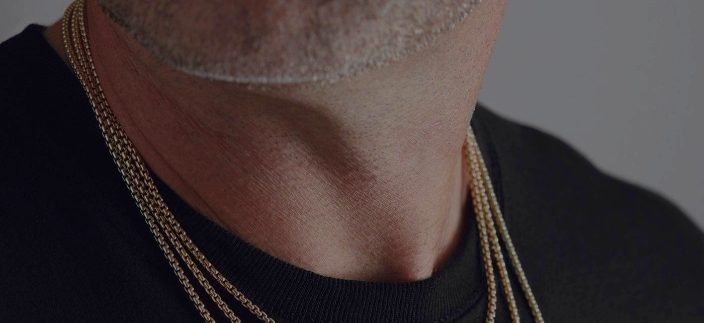 Close-up of a man's neck wearing layered gold chains over a black shirt, showcasing fine metal textures.