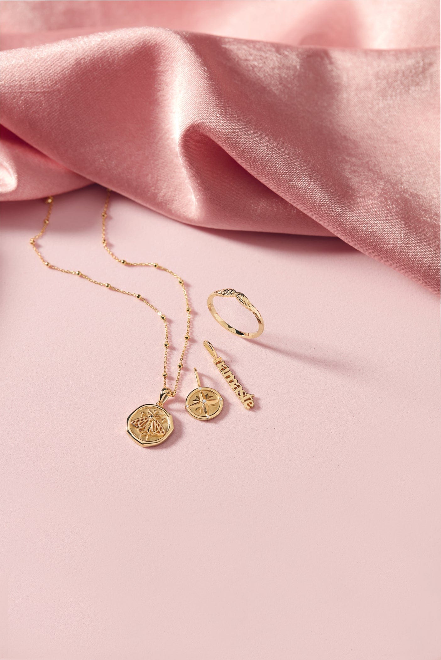Gold jewelry, including a necklace, ring, and bar pendant, on a pink surface with pink satin fabric.