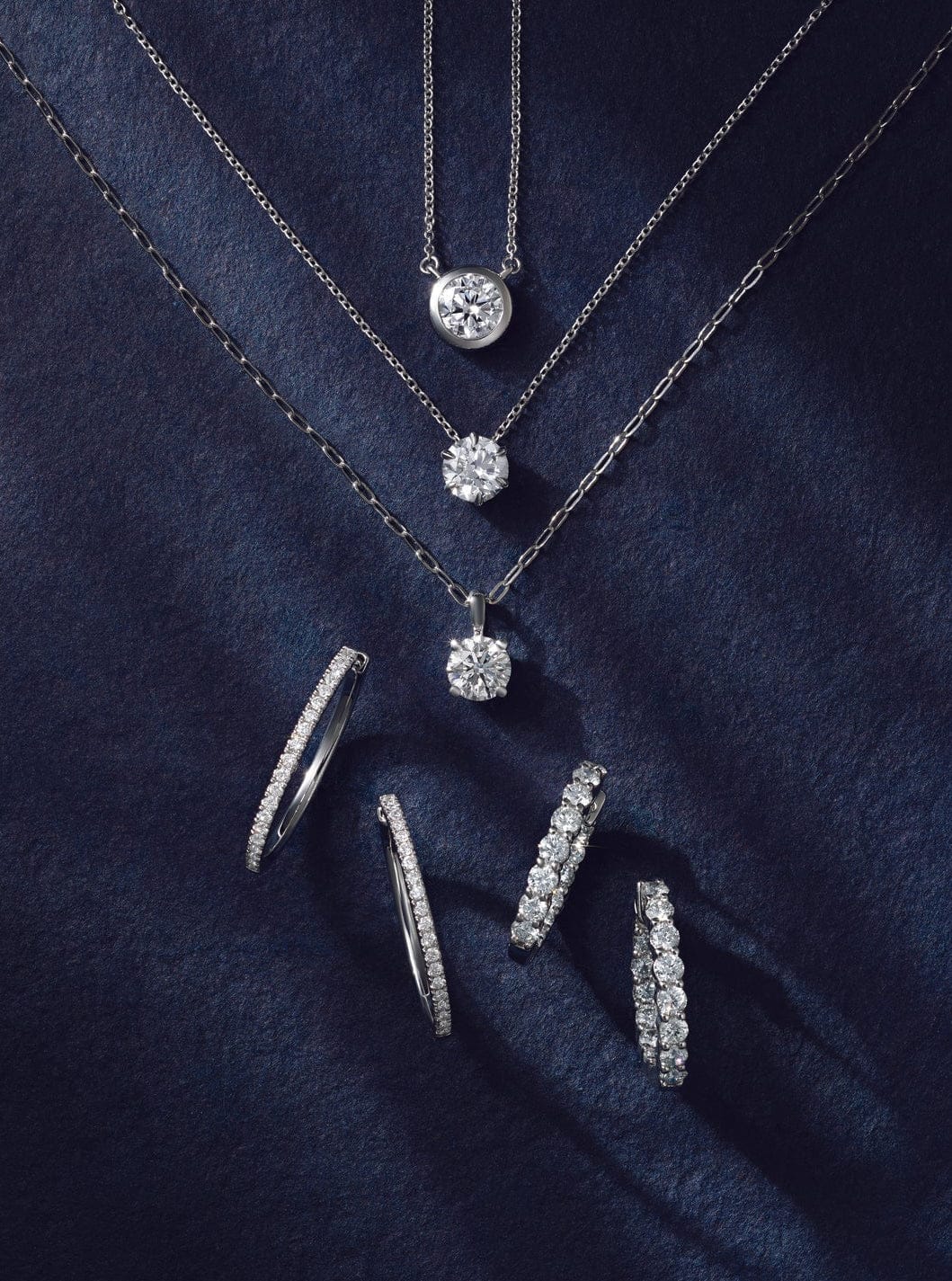 Diamond jewelry set including diamond pendant necklace, diamond hoop earrings, and ring on a dark blue fabric background