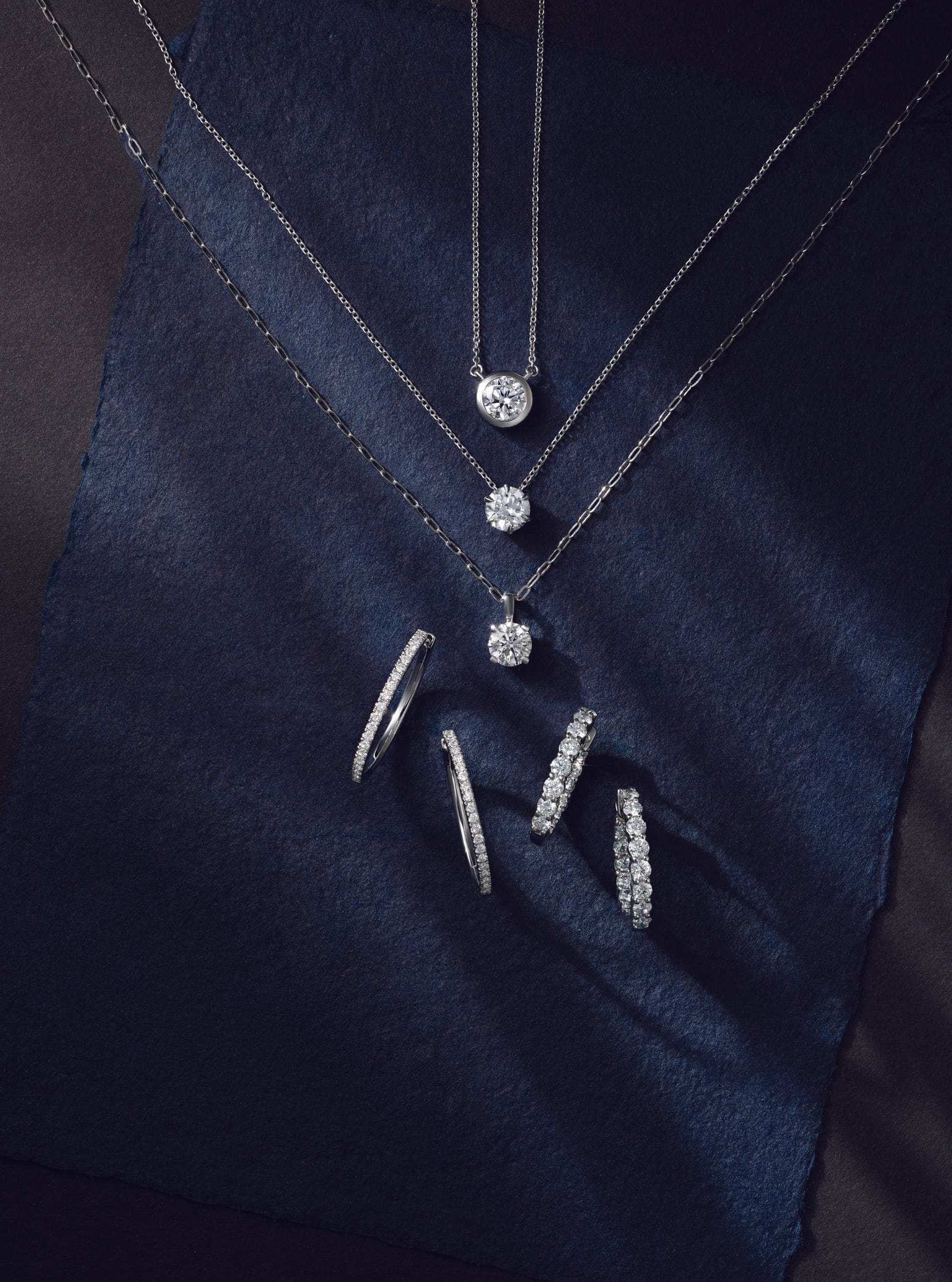 Diamond white gold pendant necklaces and diamond hoop earrngs on a dark blue fabric background