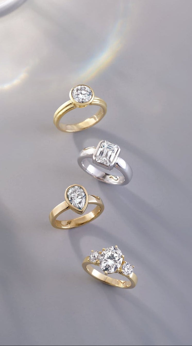 Collection of gold and silver bezel set engagement rings with lab grown diamond accents on a reflective surface.