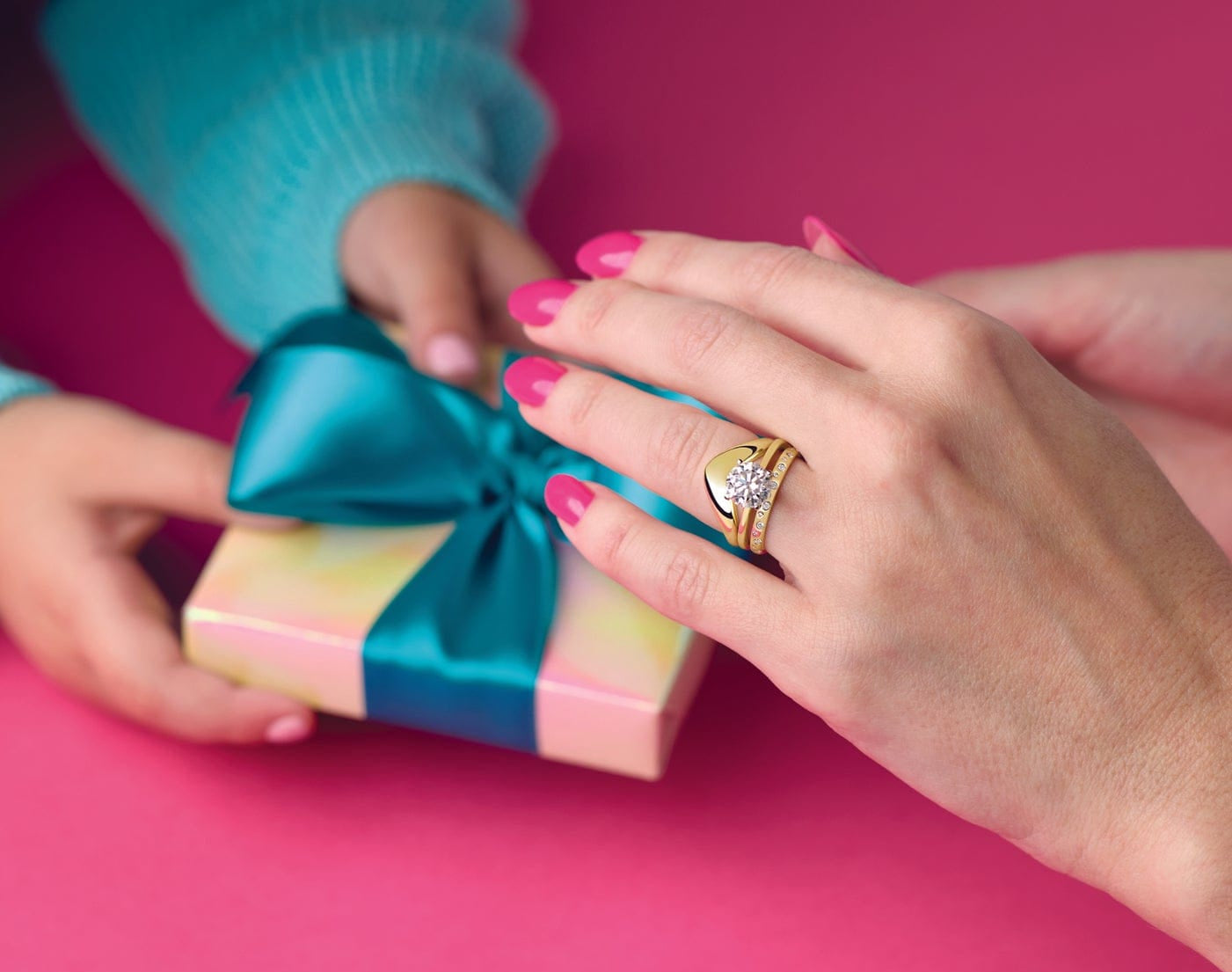 Hands exchanging a small pastel gift box tied with a teal ribbon, with one hand wearing a gold engagement ring with a round diamond.