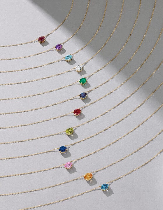 Nine gold necklaces with different colored gemstone pendants arranged in curved, parallel lines on a gray background.