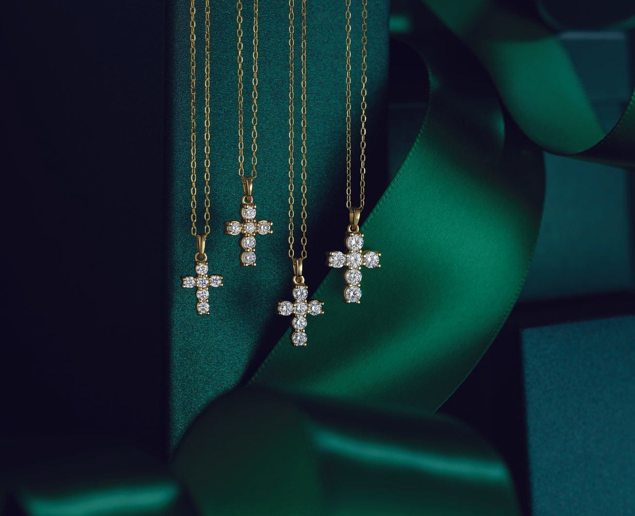 Four gold necklaces with diamond cross pendants displayed against a dark green background with ribbon.