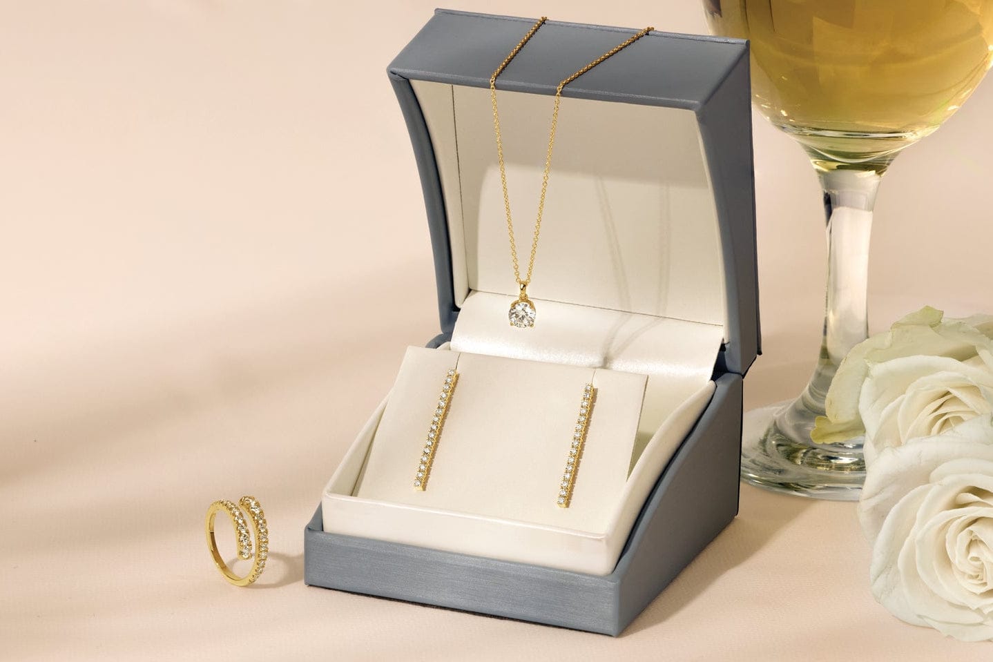 Jewelry set including diamond earrings and a ring in an open box on a table with a glass of wine and white roses.