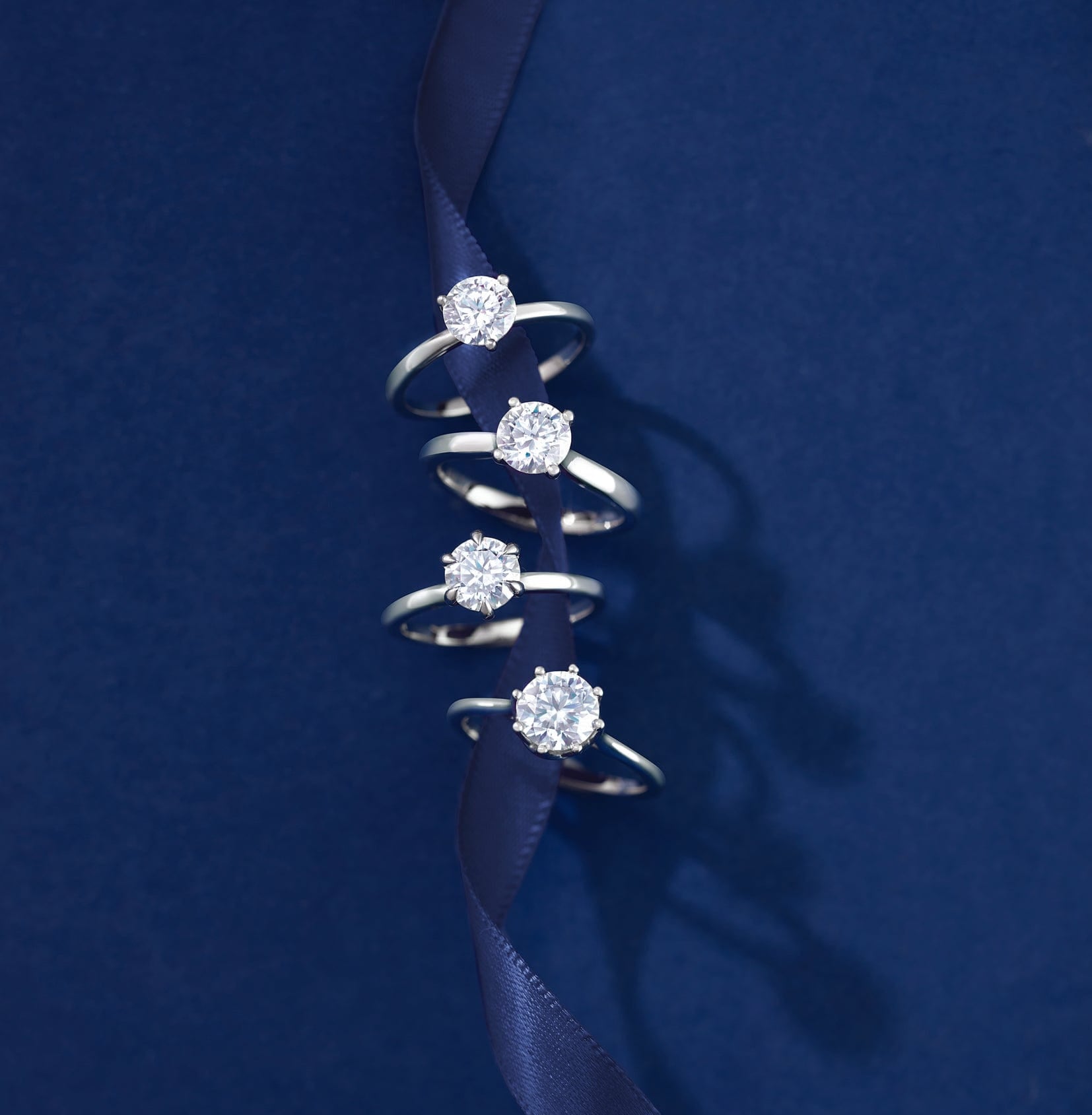 Set of 4 natural diamond solitaire engagement rings on a dark blue ribbon against a matching background