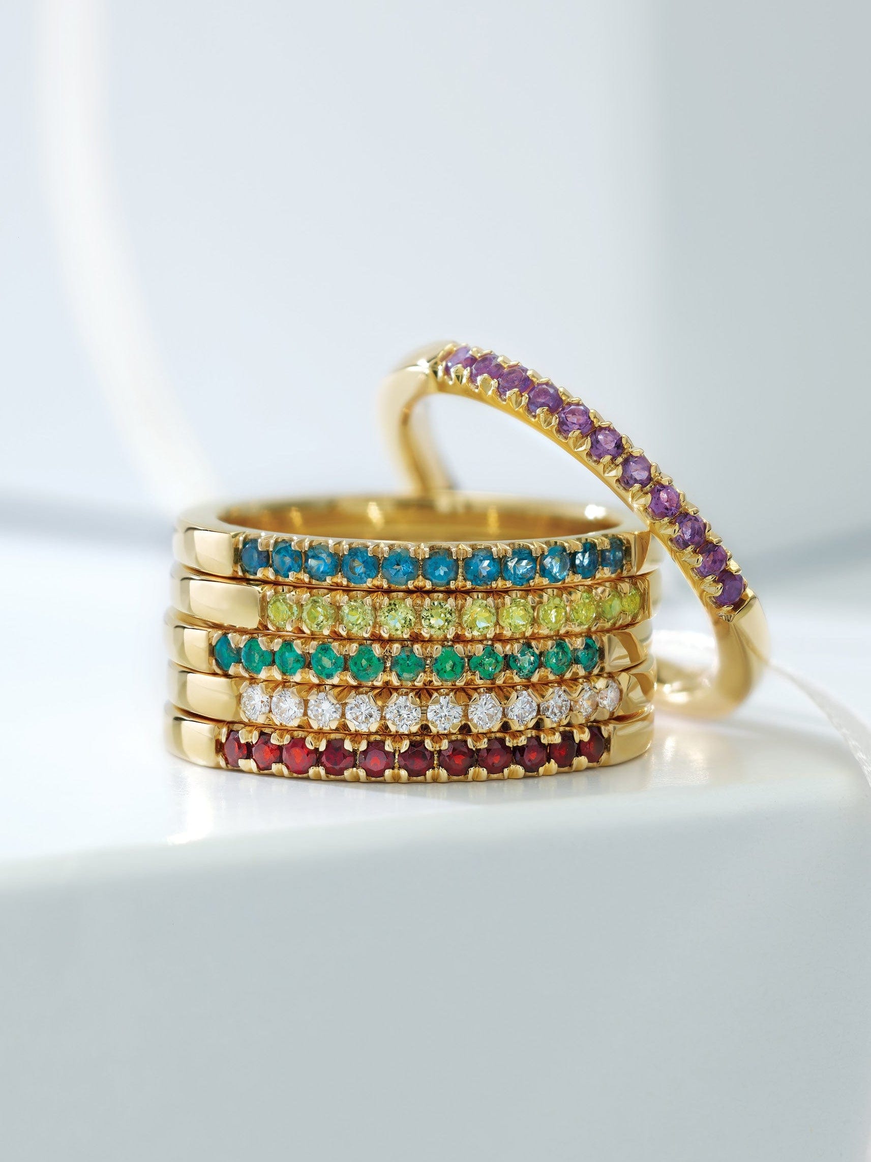 A stack of gold rings set with colorful gemstones, including blue, green, yellow, red, and purple stones.