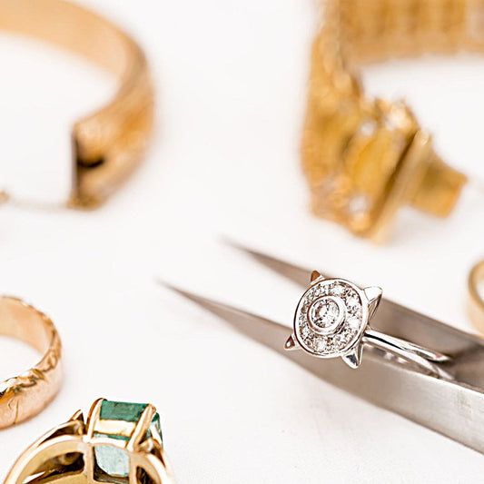 A fine jewelry repair piece, a silver ring featuring a round diamond setting that demonstrates expert restoration craftsmanship, is carefully held by tweezers amidst an array of other gold pieces on a white background.