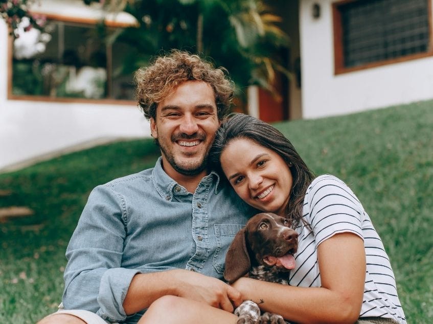 Man and woman sitting on grass with a dog, smiling at the camera.