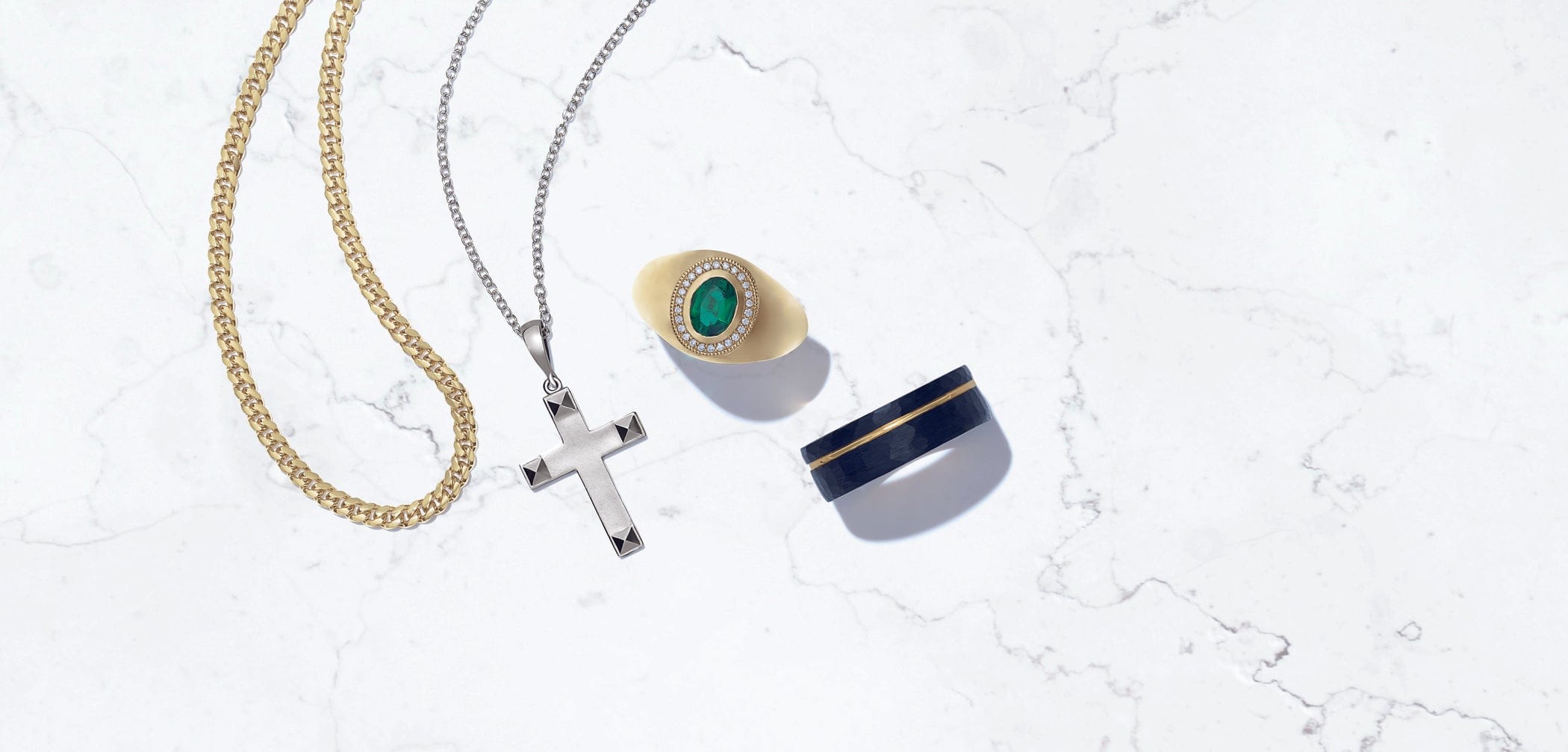 Gold chain, silver cross necklace, gold ring with green stone, and navy ring on white marble background.