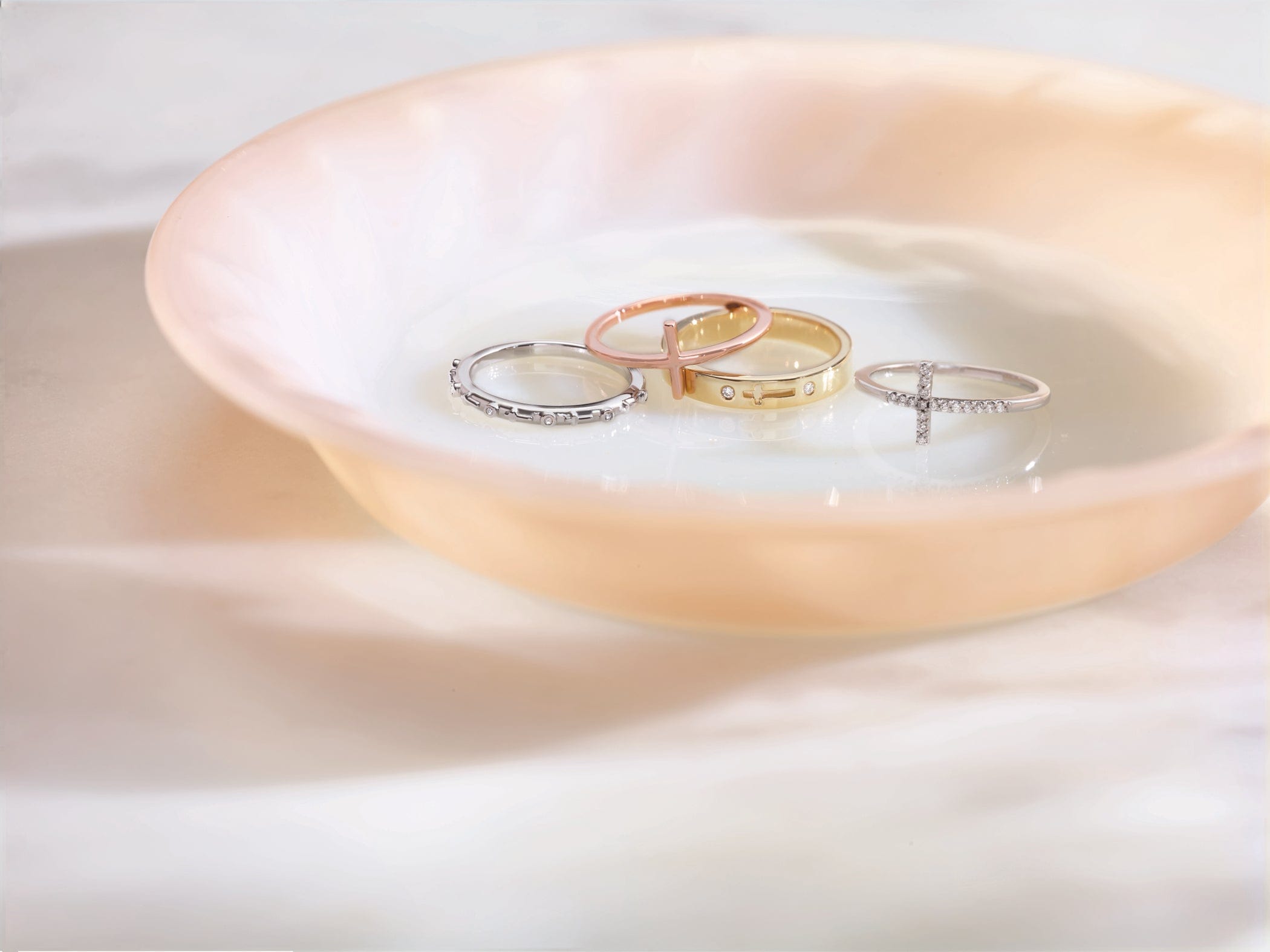 Four rings in gold, rose gold, and silver rest in a shallow, peach-colored dish on a marble surface.
