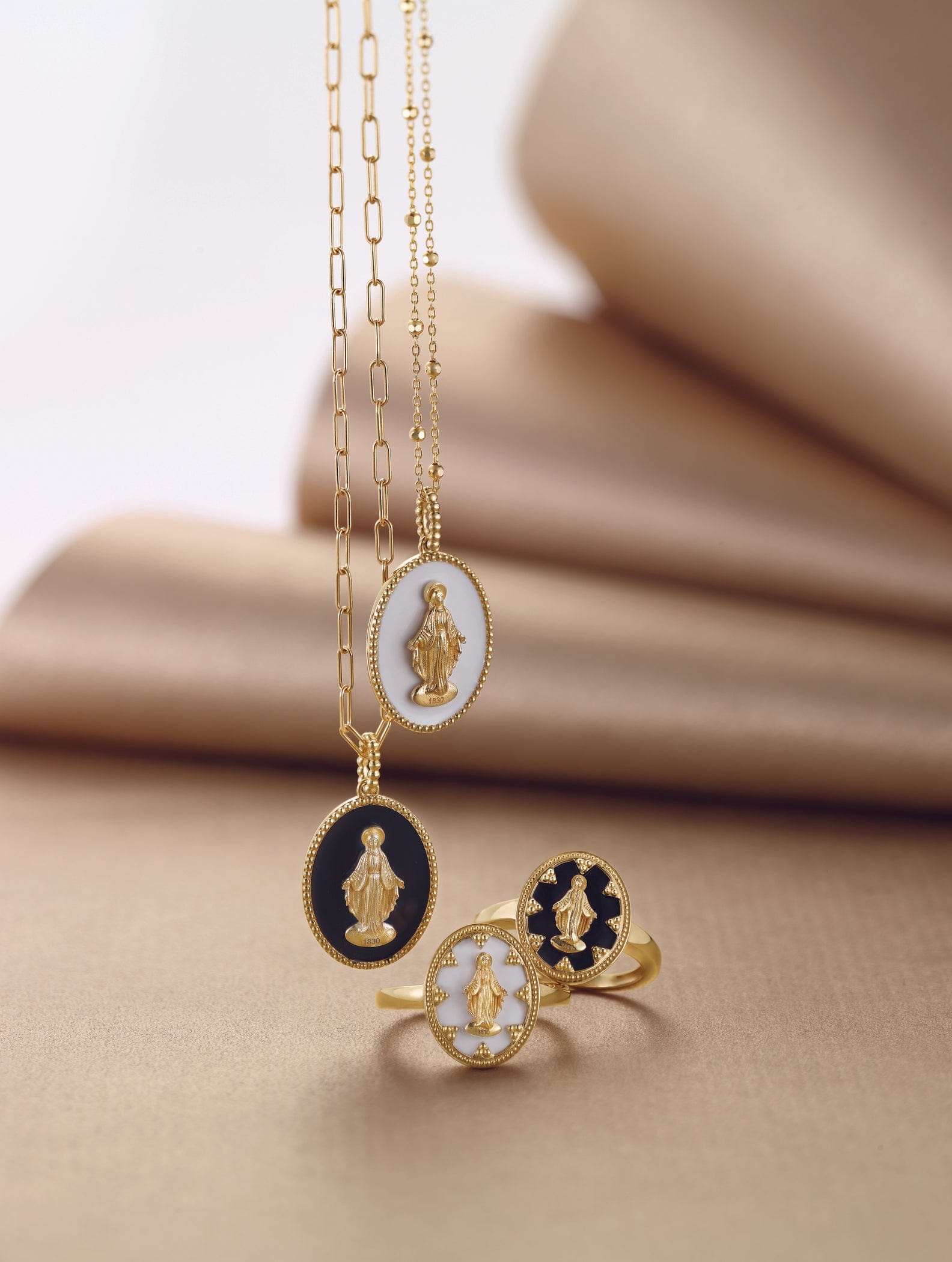 Gold necklaces and rings with oval black and white pendants featuring a gold religious figure, on a soft beige background.