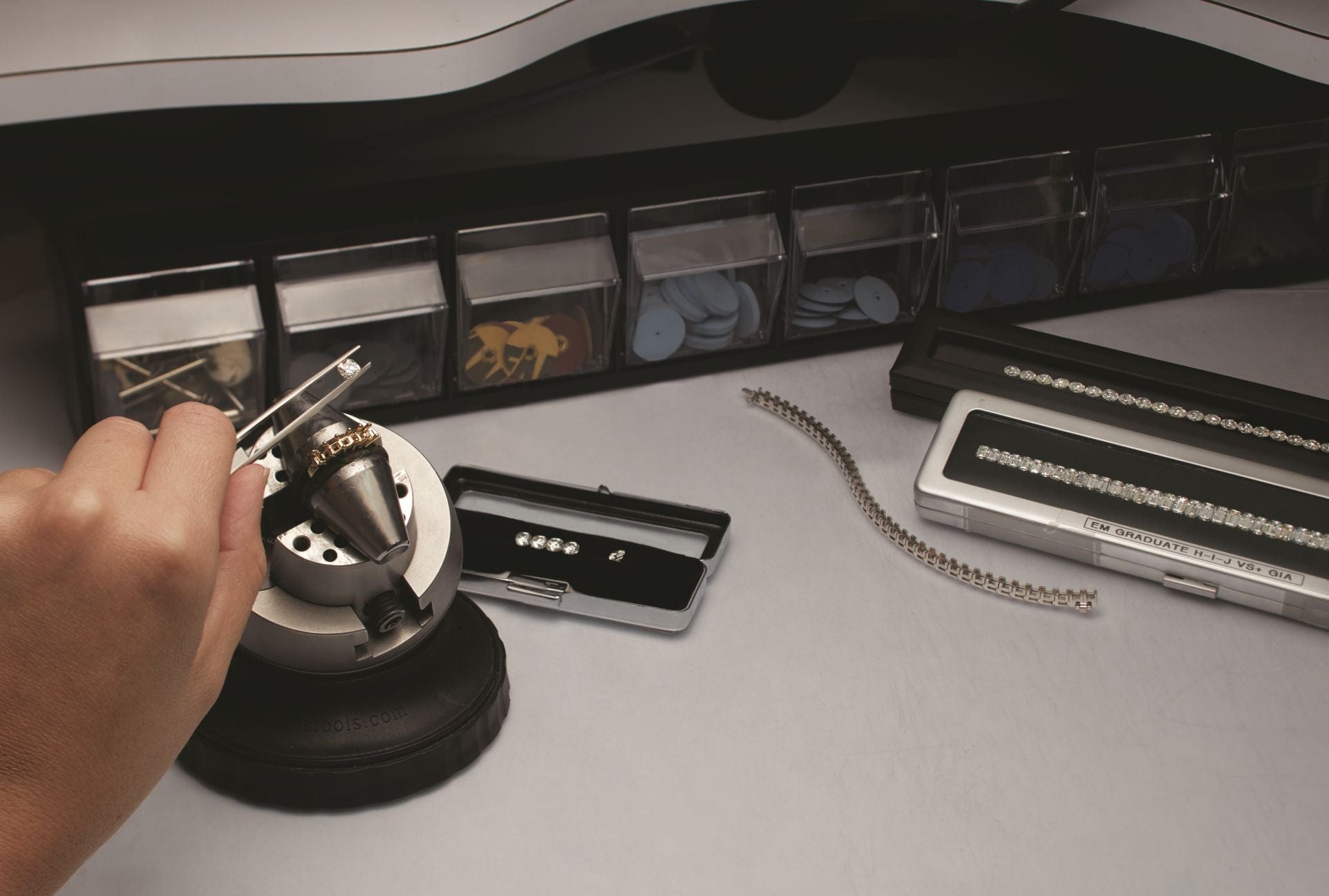 A hand holds a tool with diamonds on a workbench, with loose diamonds and tools arranged nearby.