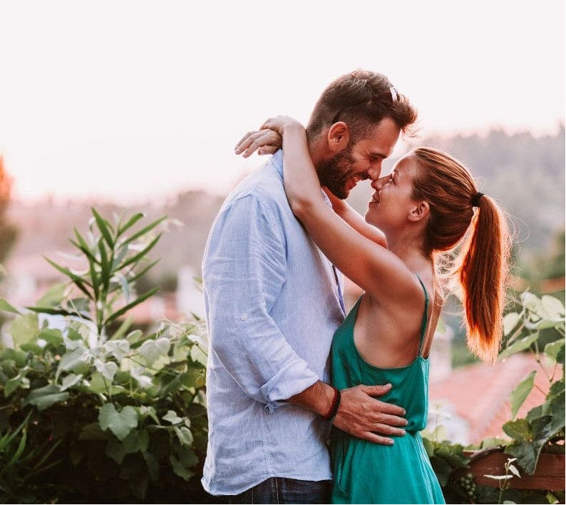 A couple in an intimate embrace outdoors, smiling warmly at each other with greenery and soft sunlight in the background.