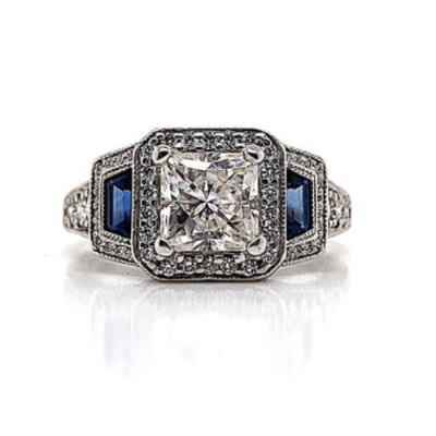 This elegant ring is crafted in 18K white gold and features a 6 mm princess-cut moissanite center stone. It is accented with .30 carats of natural trapezoid-cut blue sapphires and .60 carats total weight of natural round brilliant diamond accents, all arranged in a halo style design for timeless allure.