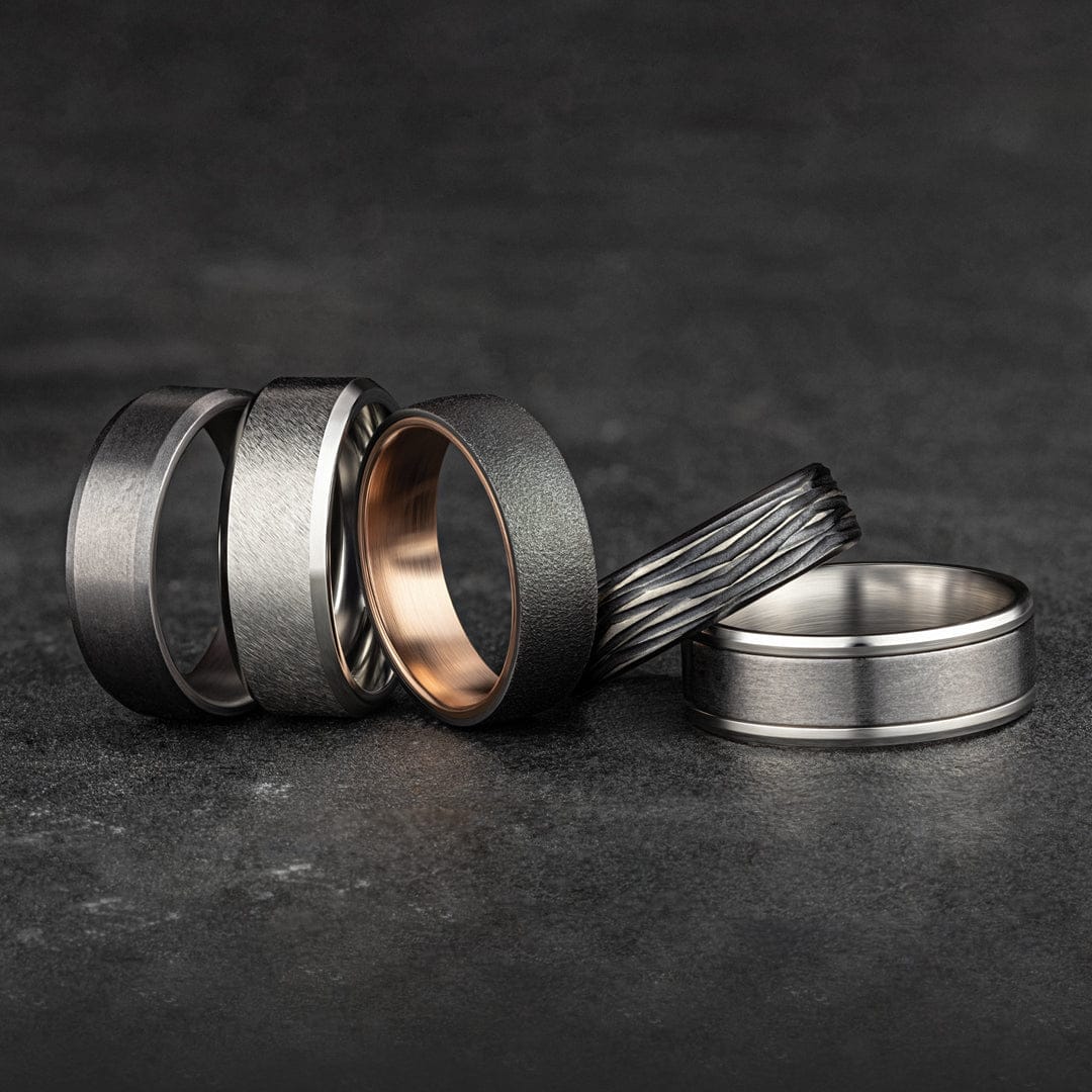Five modern wedding bands featuring varied textures and metal finishes, arranged on a dark stone surface.