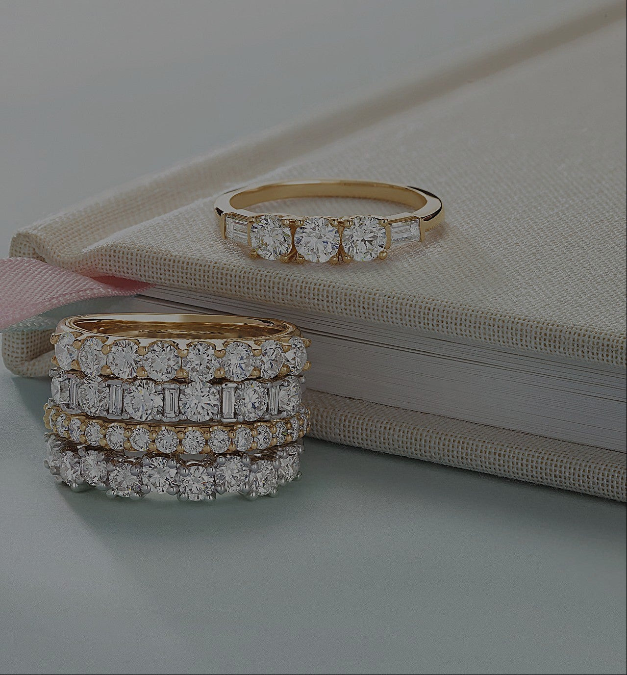 Stack of five gold and diamond rings with round and baguette cuts arranged beside a closed cloth-bound book.