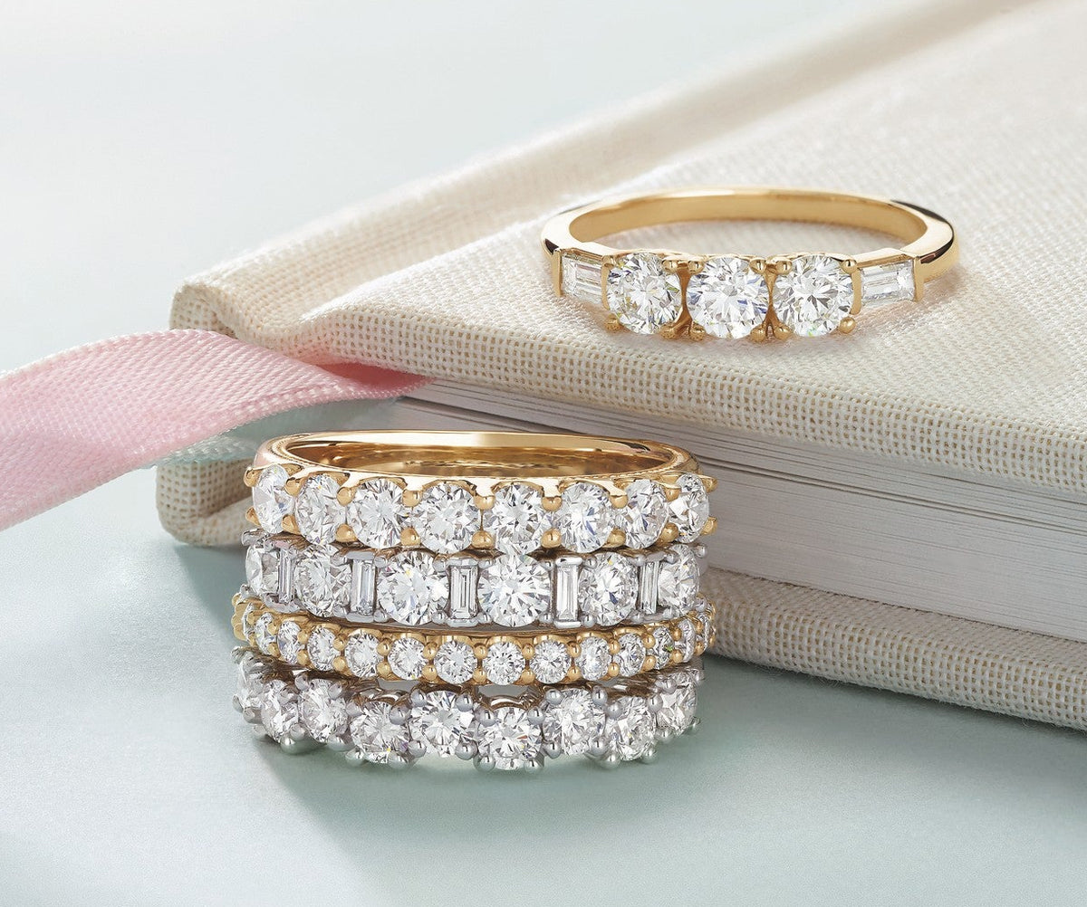 Stacked gold and silver diamond bands featuring round and baguette-cut stones displayed on a soft blue surface with a beige book and pink ribbon.