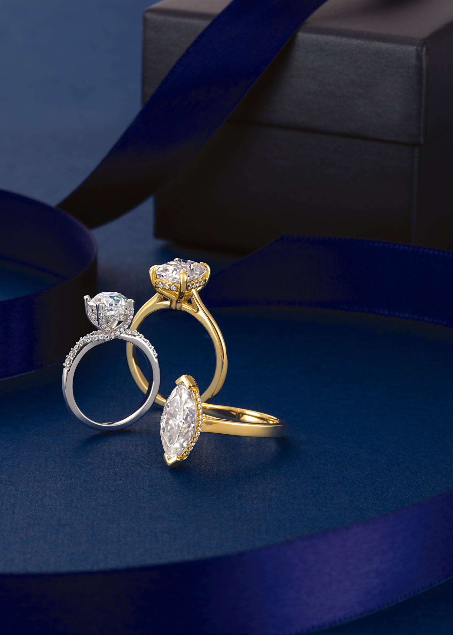 Three custom engagement rings with large marquise and round-cut diamonds set in gold and white gold bands, arranged on a deep blue surface with a matching ribbon and a dark gift box in the background.