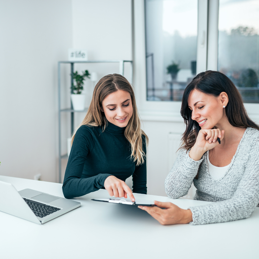 In their brightly lit office, two women are deeply immersed in a spirited discussion, gesturing towards a tablet. A laptop on the table hums with inspiration, laying the groundwork for an expert consultation through Custom Jewelry Consultation to craft one-of-a-kind jewelry designs tailored to perfection.