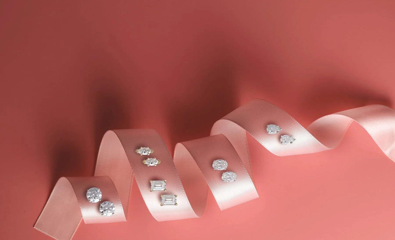Diamond stud earrings arranged on a pink ribbon against a pink background.