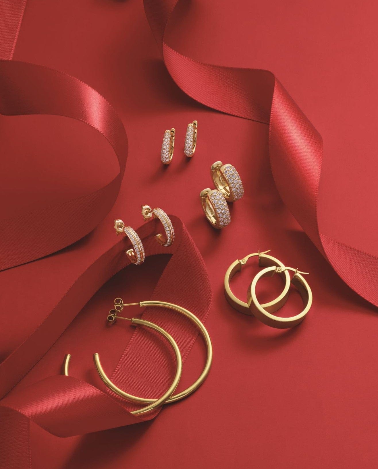 Gold hoop earrings and diamond-studded earrings arranged on a red background with red satin ribbons.