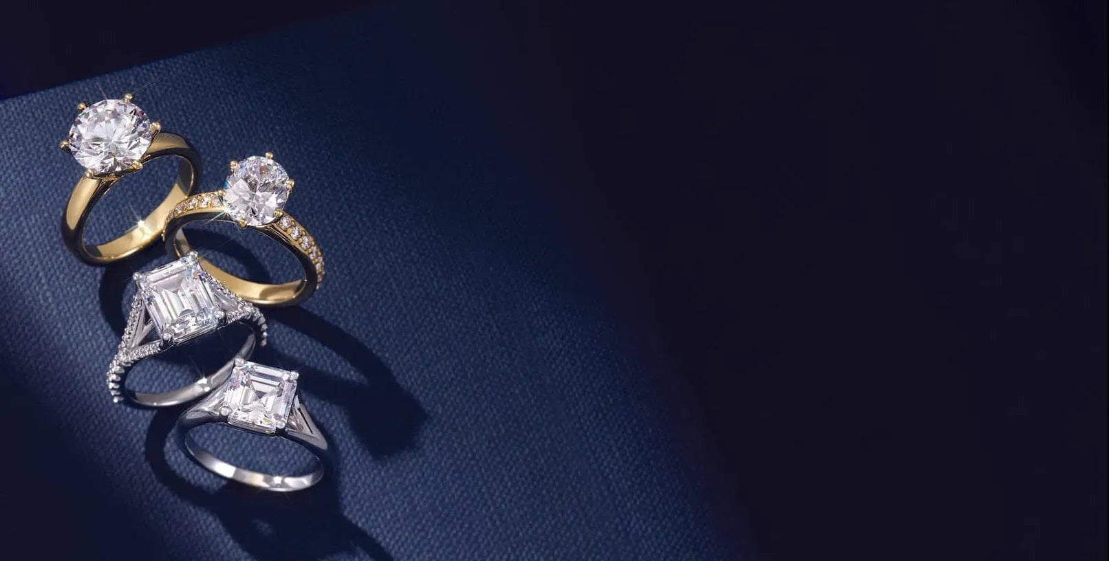 Four diamond engagement rings with gold and silver bands arranged diagonally on a dark blue surface with soft lighting highlighting the gemstones.