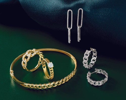 A selection of elegant gold and silver jewelry pieces including a gold chain bracelet, two gold rings with diamond accents, silver hoop earrings with diamond embellishments, and long rectangular silver drop earrings displayed on a deep green fabric background.