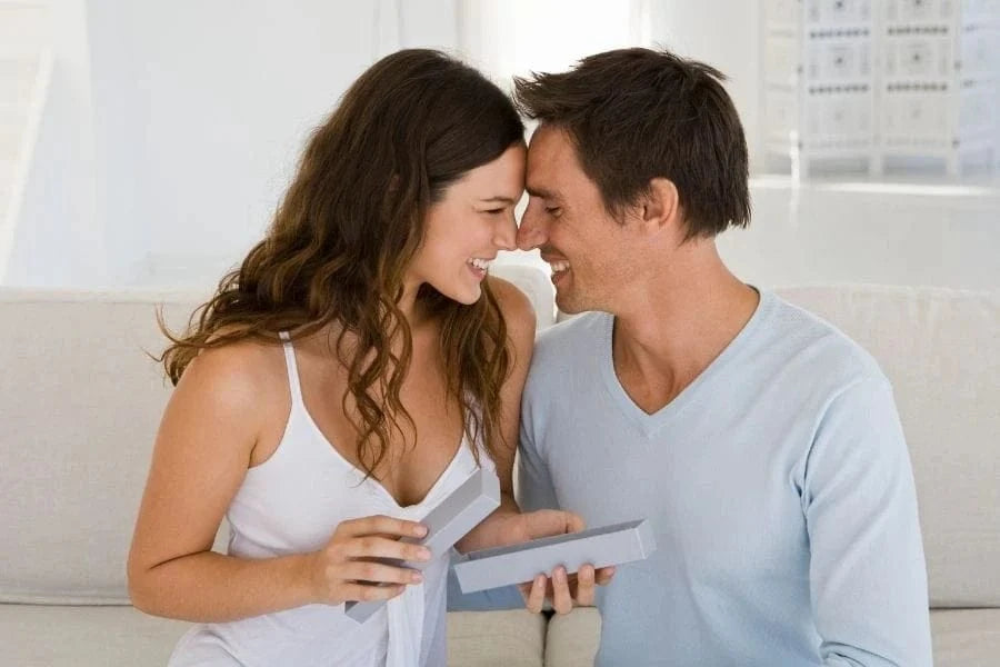 A smiling couple touches foreheads while exchanging a gift on a white couch.