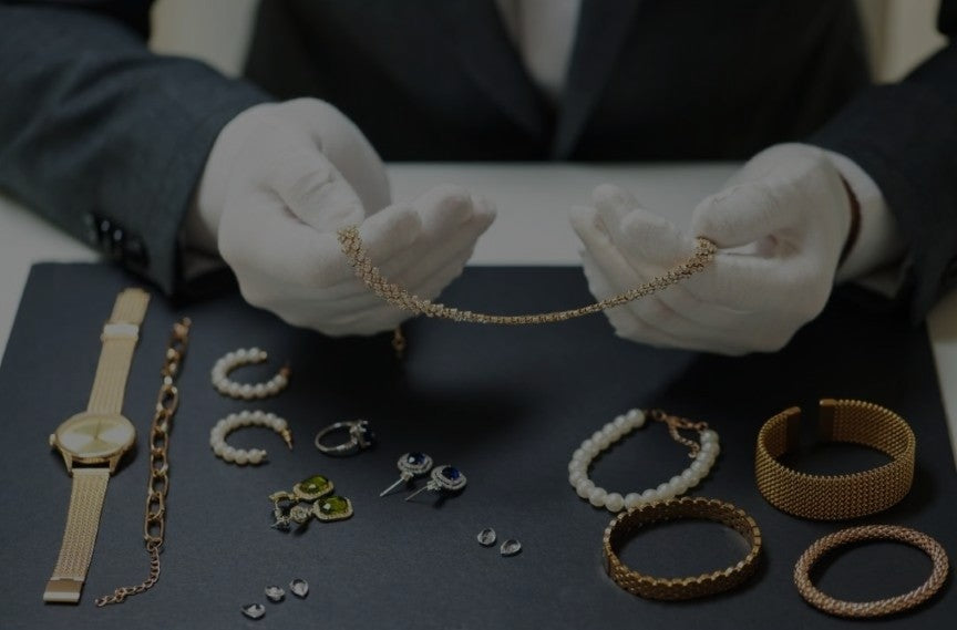 A person wearing white gloves examining a delicate gold chain necklace on a black surface surrounded by various gold and gemstone jewelry pieces including bracelets, earrings, and rings.