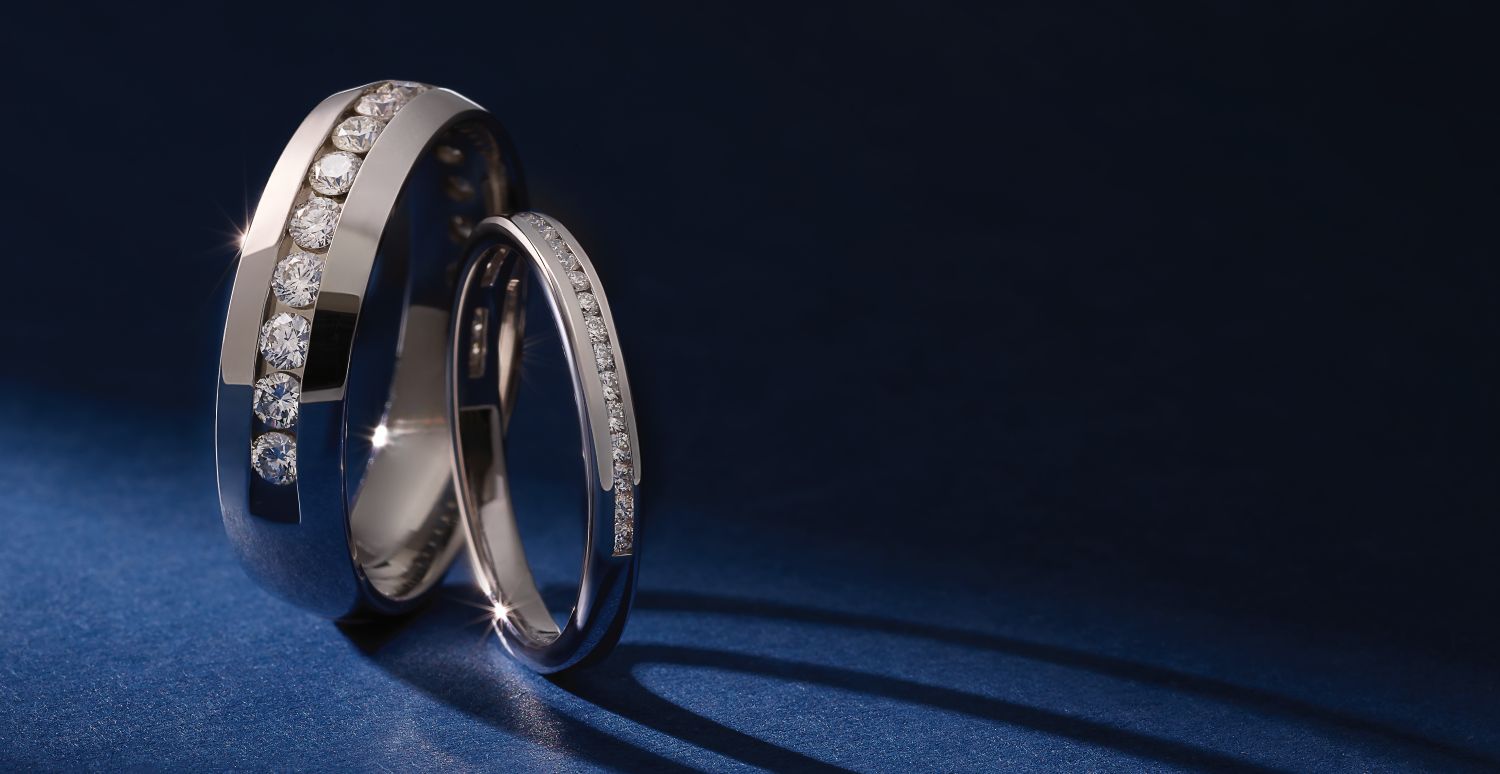Two silver wedding bands with rows of inset diamonds positioned upright on a dark blue surface casting subtle shadows.