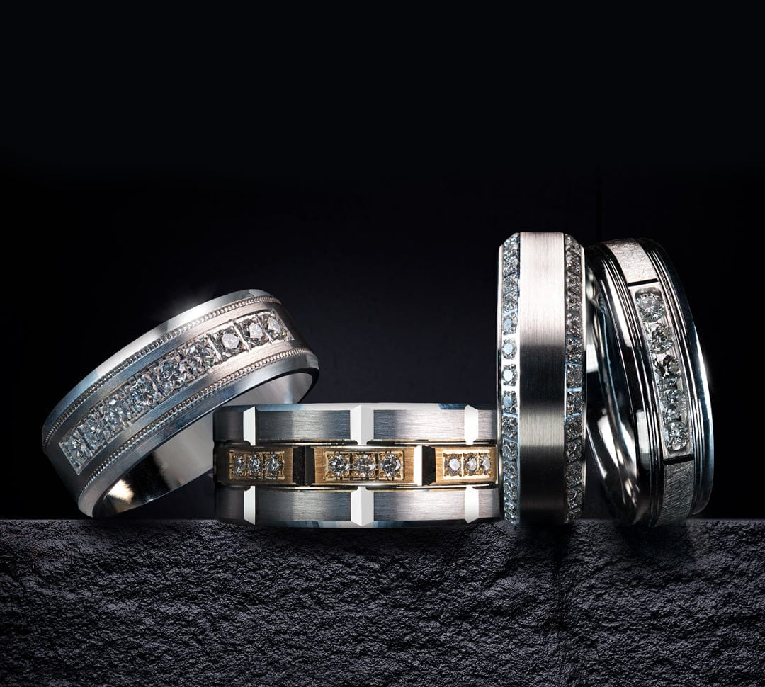 Four elegant diamond-studded mens rings in silver and gold, displayed on a dark textured surface.