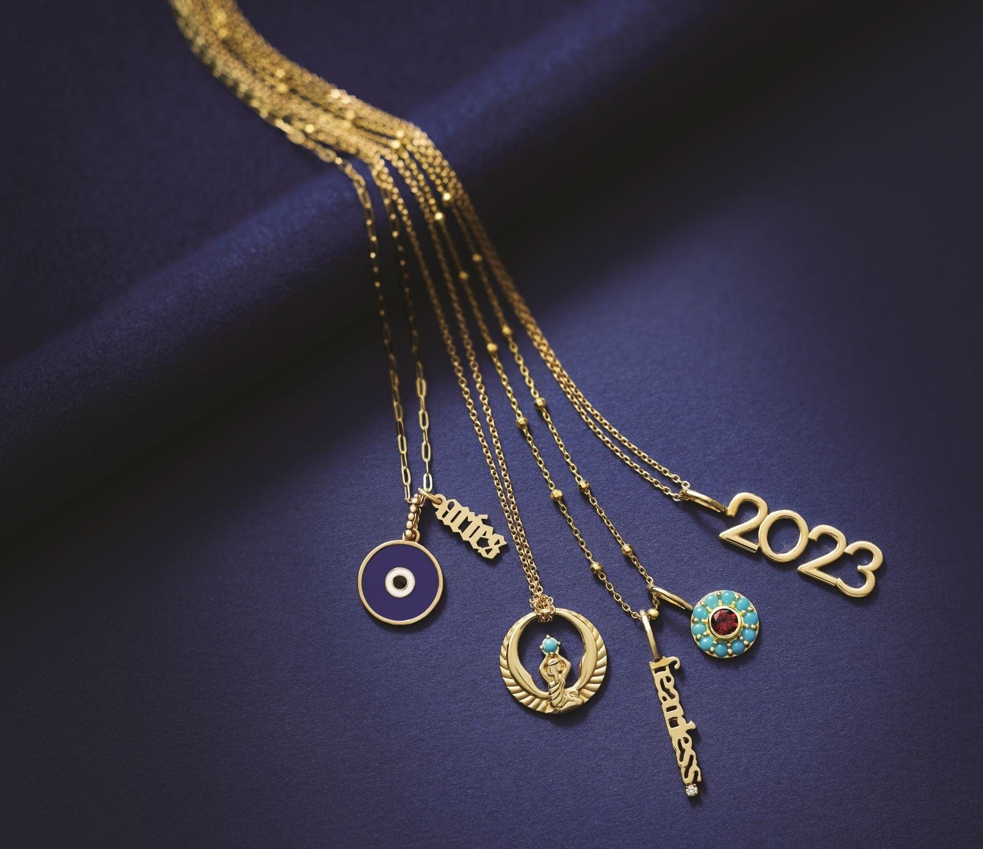 Five gold necklaces with unique pendants, including 2023, fearless, and symbols, on a dark blue background.
