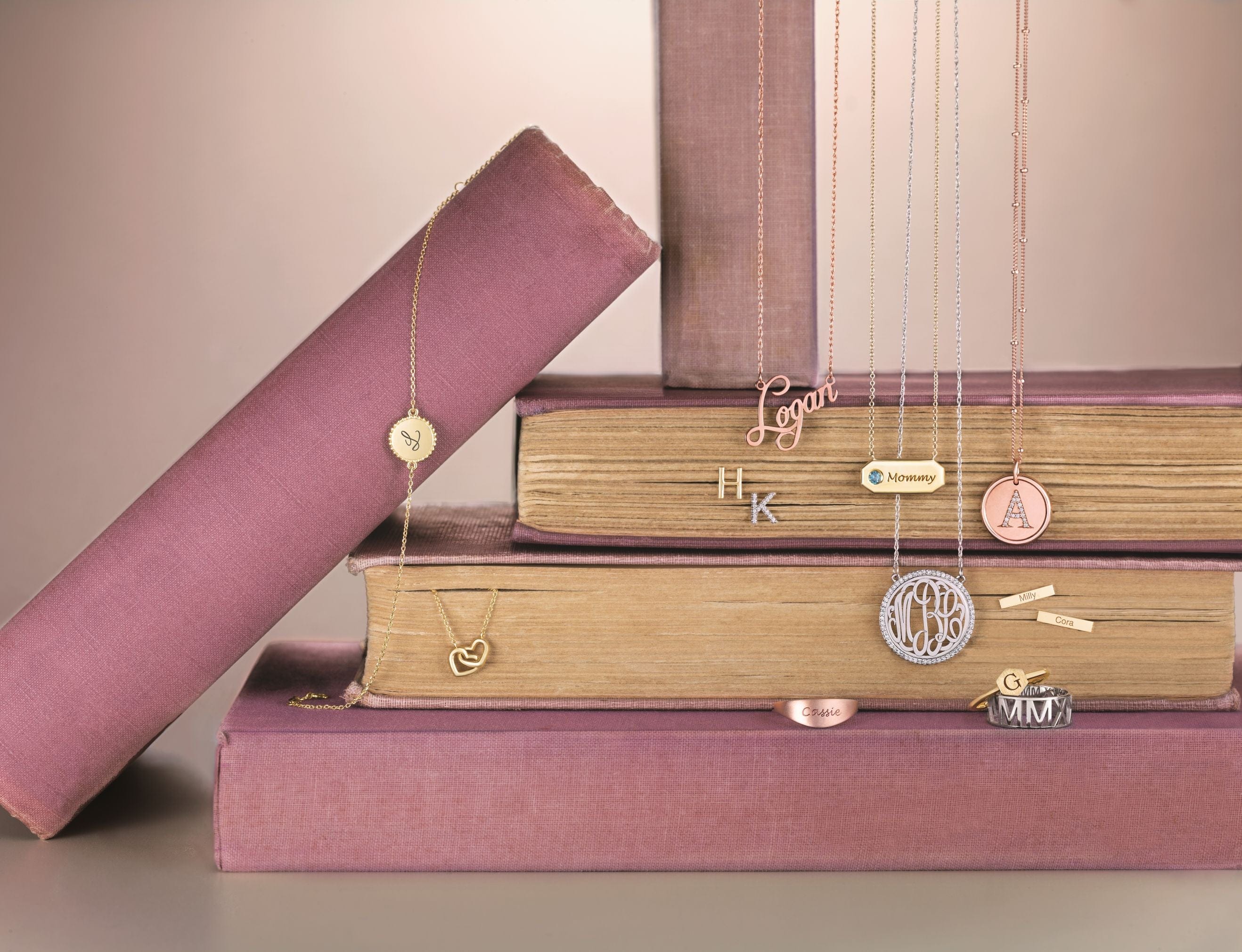 Stacked mauve and beige books displaying gold and silver necklaces with various personalized pendants.