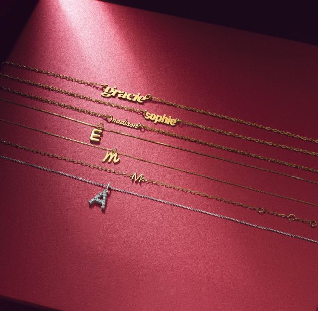 Personalized nameplate and initial necklaces in yellow gold and white gold displayed on a deep red background, featuring custom names and letter pendants with delicate chain designs.
