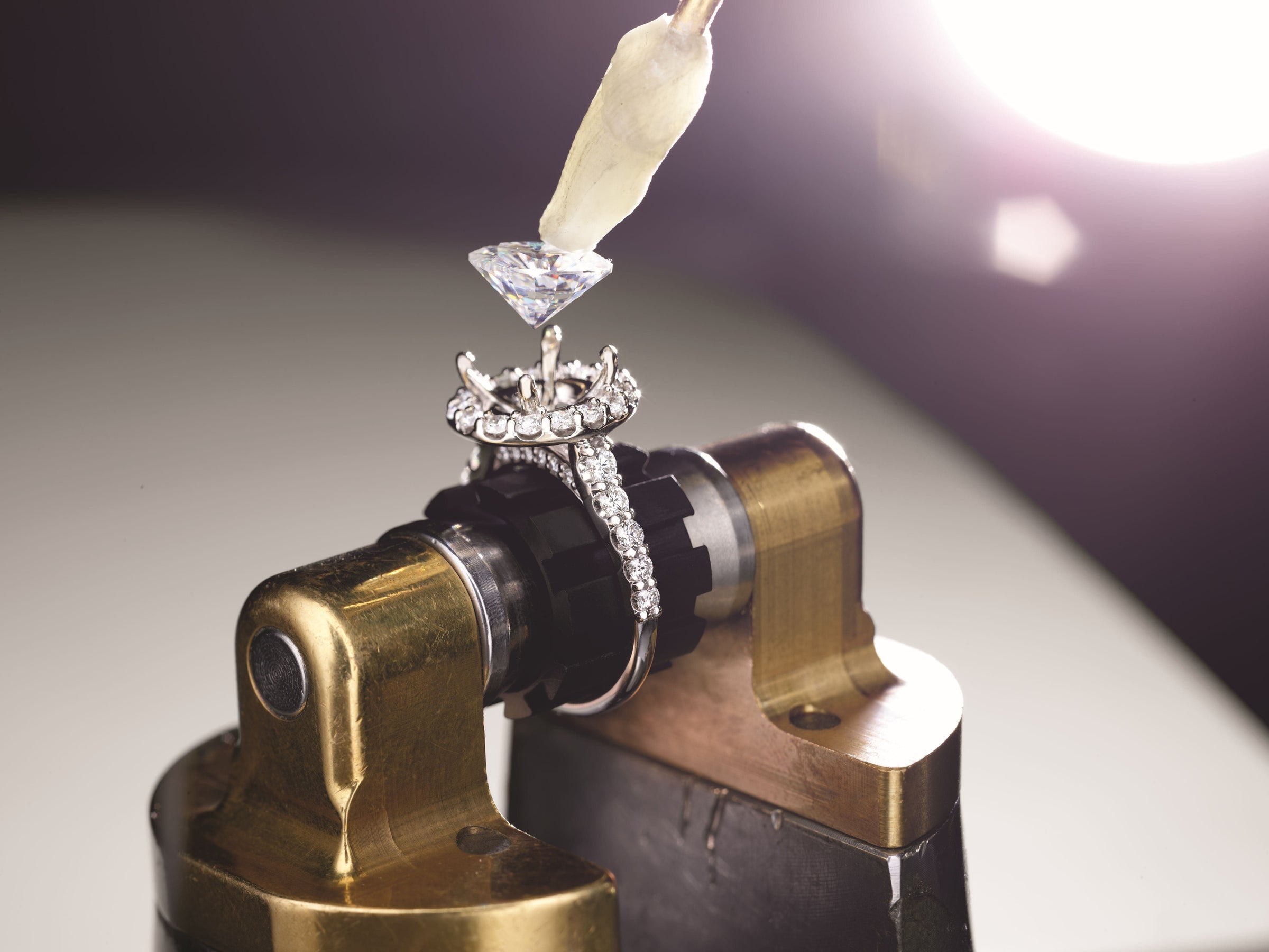 A diamond is being set into a ring held by a jewelers vise, with bright light in the background.