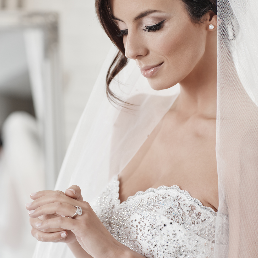 A bride, who recently had a Bridal Consultation, is elegantly dressed in a lace wedding dress, looking down with a soft smile while holding a sheer veil indoors. Her fingers are gently adorned with shimmering engagement rings.