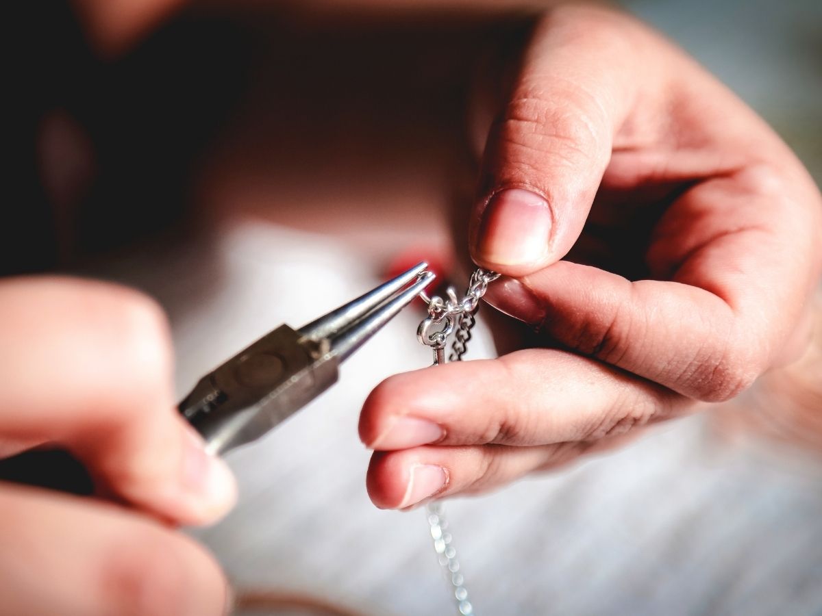 Close-up of hands using jewelry pliers to repair a silver necklace clasp with precise attention to detail.