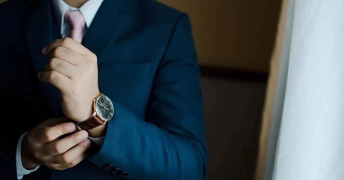 Man in a blue suit and pink tie adjusting his wristwatch near a window with white curtains.