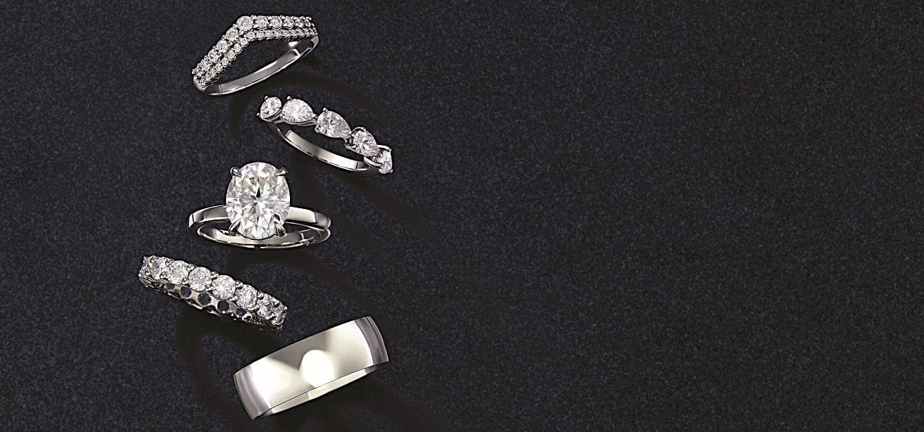 Six silver wedding bands and engagement rings displayed vertically on a dark textured surface, featuring various diamond settings and designs.