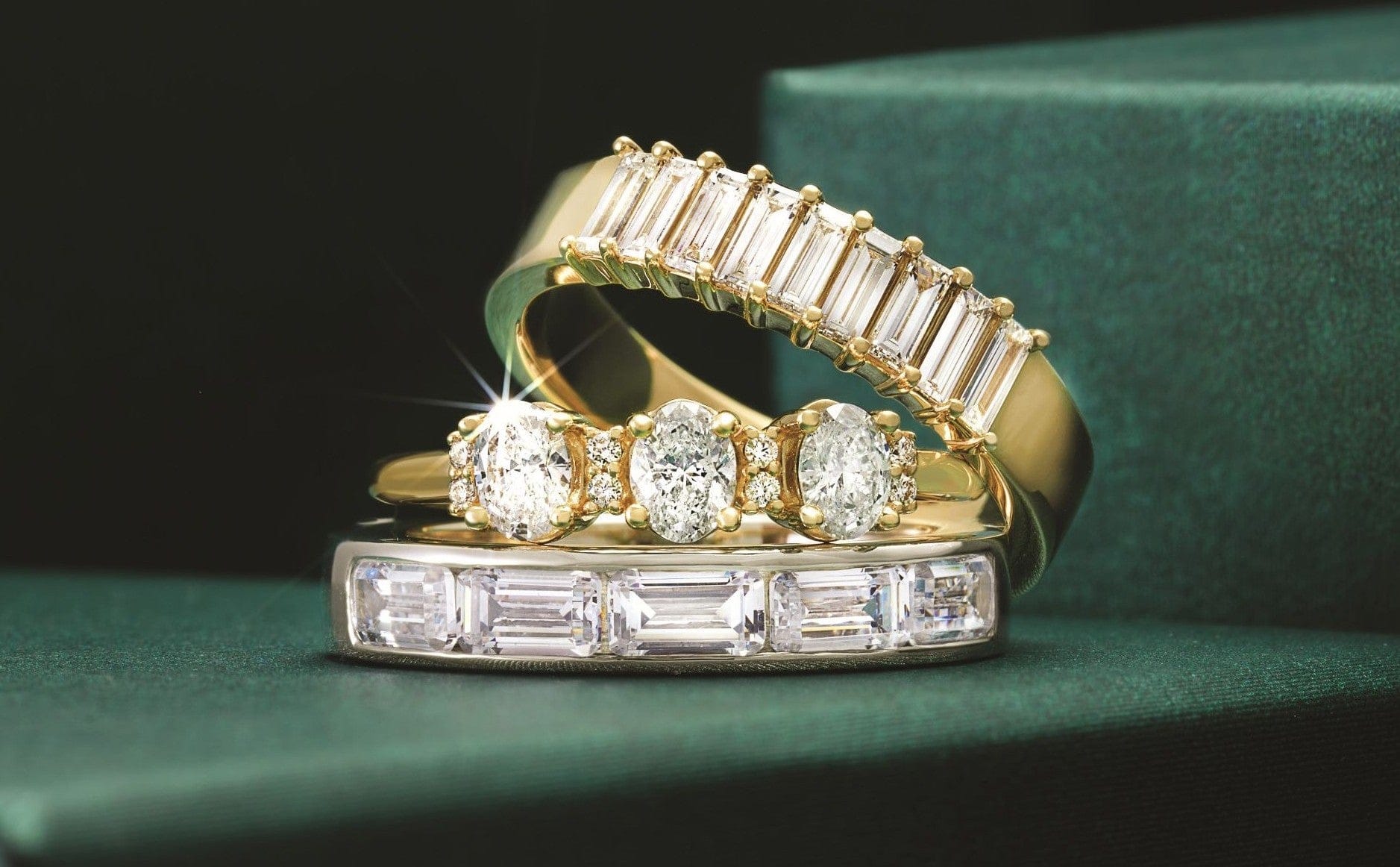 Three gold rings with sparkling diamonds displayed on a dark green surface.